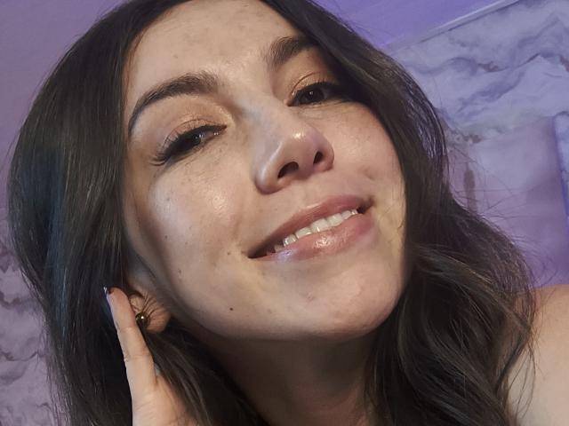 MiaReed's face with shiny skin, brown eyes, and a subtle smile, showing her teeth and slightly parted pink lips. She's touching her ear with a black-nail-painted finger.