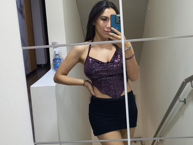 MiaReed with long dark hair, wearing a shiny purple top with small boobs and a black mini-skirt, taking a mirror selfie, showing a hand on her hip.