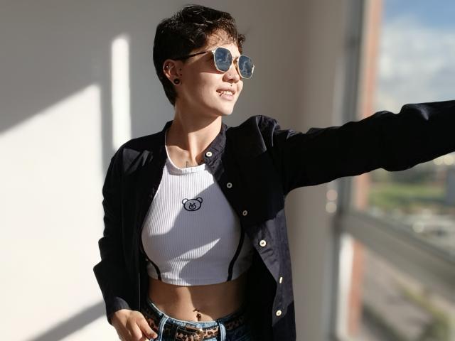 LissyLuxe with short dark hair, wearing aviator sunglasses, a white ribbed crop top, and a black button-up shirt, smiling while reaching out. Her tits are small, and she's cute.