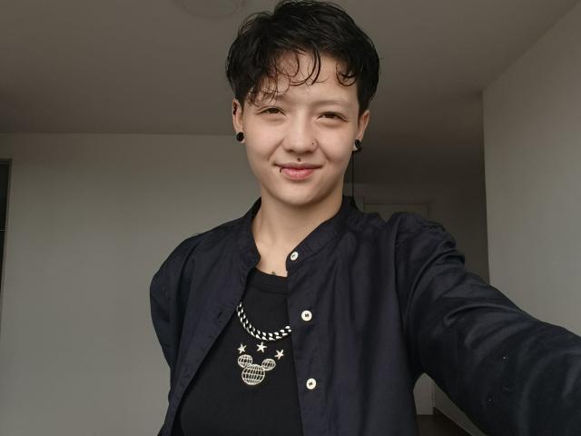 LissyLuxe, capelli neri corti, camicia nera, collana con teschio, espansori per le orecchie neri, sorridente, lingua leggermente fuori, pelle media, piccolo piercing al naso, giacca blu scuro, selfie..