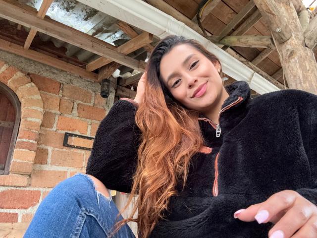 MyaSpecter with long, wavy, brown hair, smiling, wearing a black fleece zip-up and ripped blue jeans, her hand in her hair, showing pink nails, under a wooden roof.