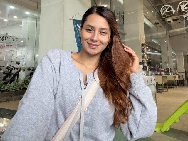 MyaSpecter with long brown hair, grey hoodie, and pink lips, smiling slightly, hand in hair, beige strap across chest, indoor setting, casual vibe.