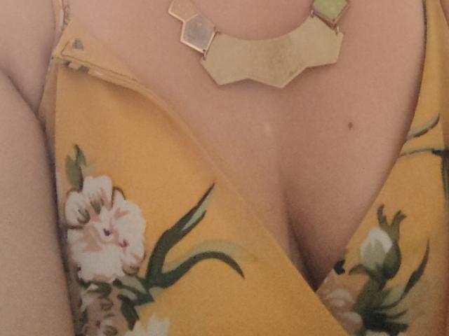 O vestido floral amarelo de VioletaSwitch, que mostra seus seios em um decote profundo, com um colar geométrico dourado repousando em seu peito. Sua pele clara tem uma pequena pinta..