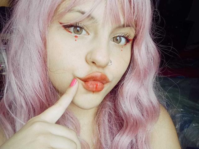 Sophie Hill, com cabelo rosa, piercing no nariz e delineador vermelho, fazendo beicinho com o dedo nos lábios, batom vermelho, unhas rosa e pequenas manchas vermelhas na bochecha..