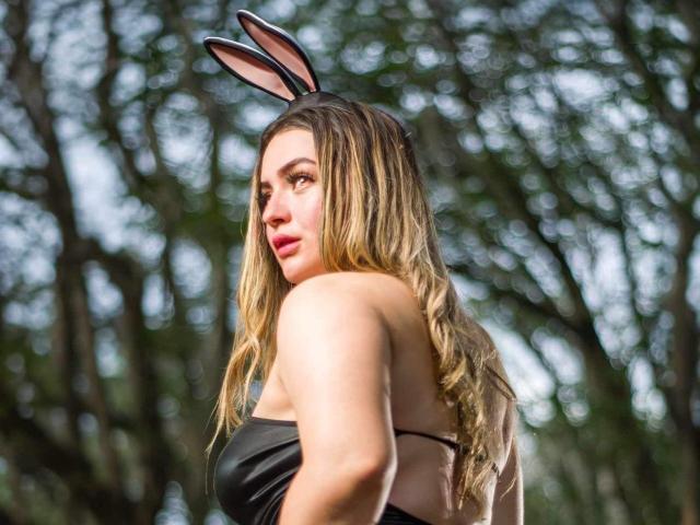 GigiHeart69 met lang, golvend blond haar, zwarte konijnenoren en rode lippenstift, draagt een zwarte top zonder bandjes en kijkt over haar schouder, waarbij haar blote rug en borsten zichtbaar zijn..