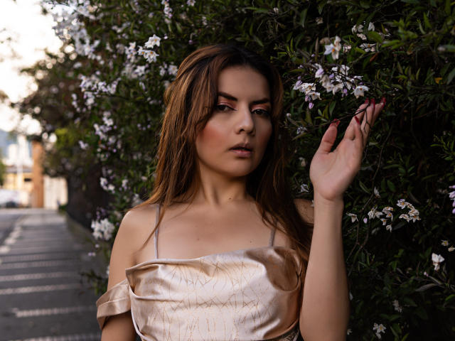 MandyMouses with wavy brown hair, wearing a shiny off-shoulder top, touching white flowers in greenery, has a serious expression, showing her red-painted nails.