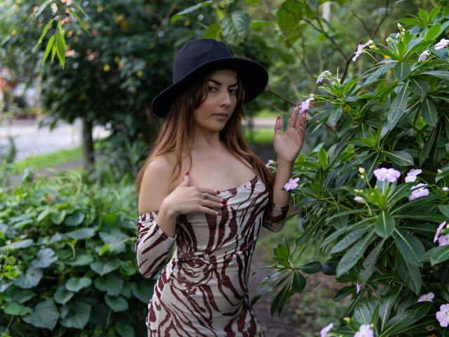MandyMouses met bruin haar, die een zwarte hoed en een jurk met bruine strepen draagt, met een open schouder, die roze bloemen aanraakt, waarbij haar borsten zichtbaar zijn, staand in een groene, bladerrijke tuin..