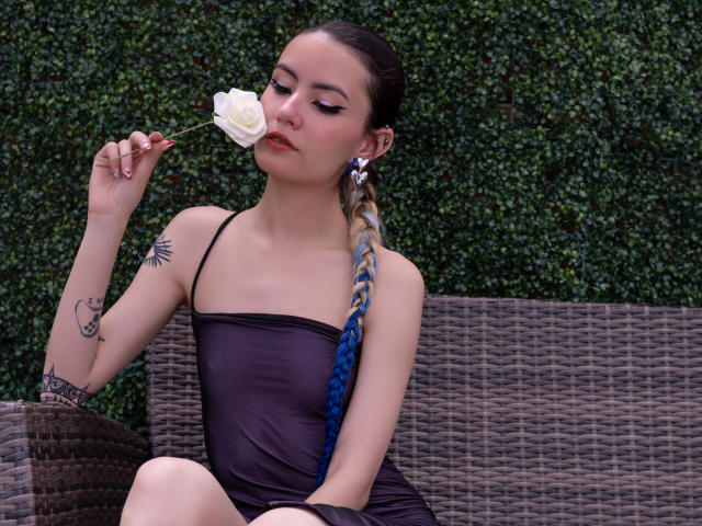 LilaLaurent con una rosa bianca in bocca, che indossa un abito nero trasparente, seduta su una panca di vimini, con una treccia blu, tatuaggi sul braccio e sulla spalla, sfondo con foglie verdi..