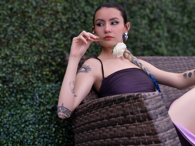 LilaLaurent with tattoos, dark brown hair in a braid, black top, holding a white rose, sitting in a wicker chair, greenery background, looking at the camera with a slight smile.