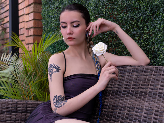LilaLaurent con tatuajes, blusa marrón oscuro, sosteniendo una rosa blanca, cabello azul trenzado, sentada en un sofá de mimbre, mirando hacia abajo, uñas pintadas de rojo, pared de ladrillo y vegetación en el fondo..