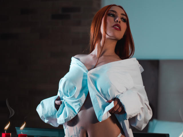 KarlyAngeel, avec ses cheveux roux, chemise blanche à épaules dénudées, laissant entrevoir ses seins et ses tatouages, tenant la chemise ouverte, yeux bleus, rouge à lèvres rouge, bougies en arrière-plan, sexy, confiante, séduisante..