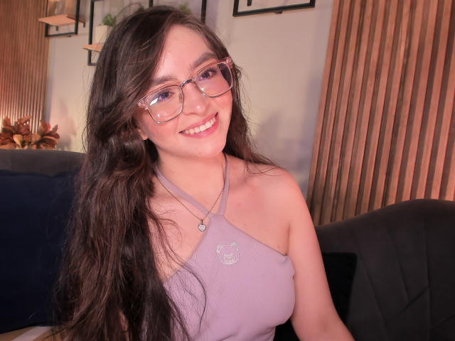 EvaMcAllister, avec de longs cheveux bruns, souriante, portant des lunettes et un haut violet à encolure dégagée, montrant ses seins, assise sur un canapé bleu foncé, avec un mur en bois en arrière-plan..