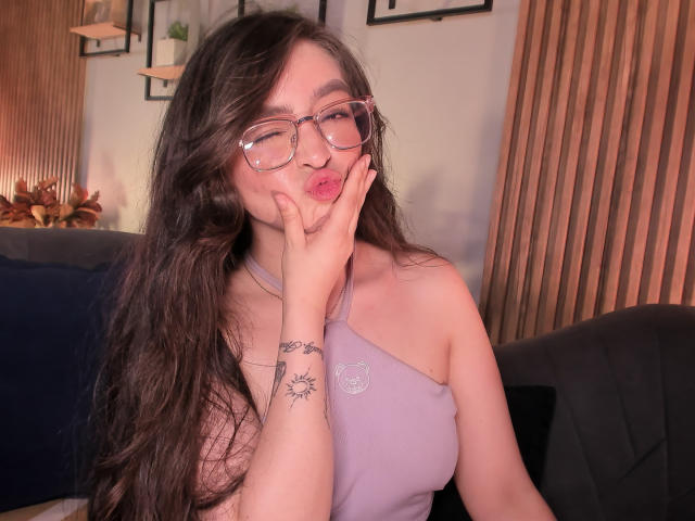 Eva McAllister, con cabello castaño largo, gafas y una blusa morada con un solo hombro, haciendo pucheros con la mano en la mejilla, mostrando un tatuaje de sol en la muñeca. Se ven los senos..