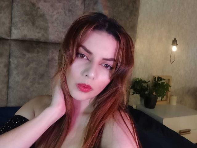 Ayneboudoir met rood haar, een bril en een glanzend mouwloos topje staat voor een spiegel, waarbij haar zwarte rok, kniehoge laarzen en reflectie te zien zijn. Ze kijkt naar zichzelf..