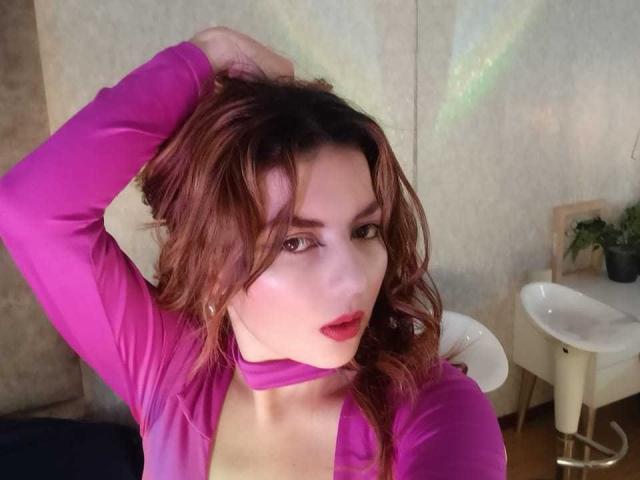 HanaGardinier, met lang bruin haar, volle lippen en subtiele make-up, draagt een donkere top en kijkt opzij met een licht geopende mond in een zwak verlichte kamer..