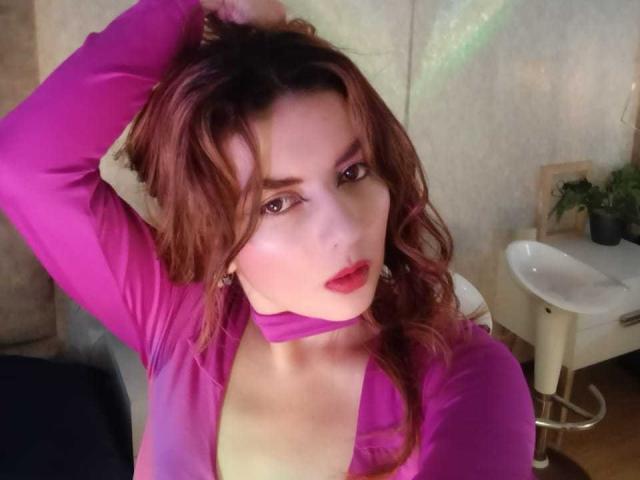 HanaGardinier, met bruin haar, groene ogen en een lichte glimlach, die haar roze lippen aanraakt met een vinger, ziet er schattig en bedachtzaam uit..