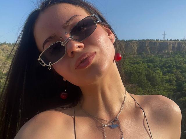 BrandyCrow with dark hair, wearing sunglasses, red earrings, and a silver necklace with a butterfly, has shiny skin and is pouting, showing her nose ring and cute face.