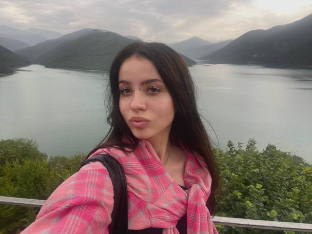 BrandyCrow with long dark hair, pink plaid shirt, and black backpack, showing off her nose ring, standing in front of a lake with mountains in the background.