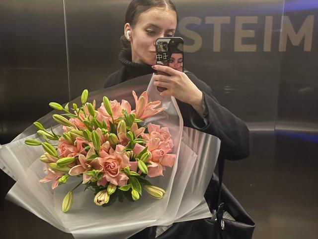 BrandyCrow with black hair in a bun, holding a bouquet of pink roses and green lilies, taking a mirror selfie in a dark, reflective room.