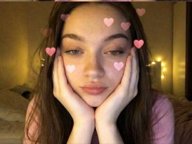 Diantrisy with hands on cheeks, pink heart emojis on face, brown hair, pink shirt, soft lighting, slightly pouty lips, bedroom background, warm yellow lights, relaxed expression.