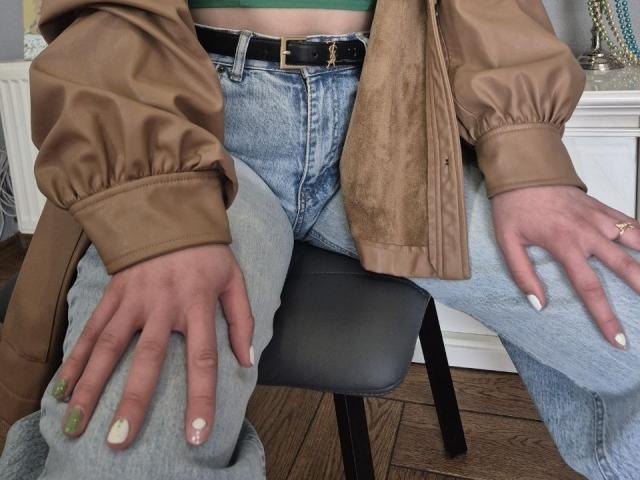 Las manos de LoriEla, con uñas pintadas, descansan sobre sus jeans, vistiendo una chaqueta marrón, un cinturón negro y una blusa verde, sentada en un taburete gris, con suelo de madera..
