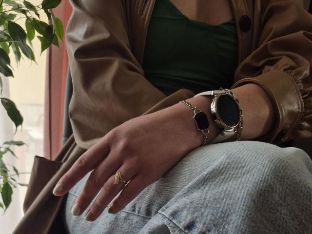 LoriEla's Hand mit einem goldenen Ring, einer schwarzen Smartwatch, einem grünen Oberteil, Jeans, einer braunen Lederjacke, sitzend in der Nähe einer Pflanze. Ihre Nägel sind gelb lackiert..