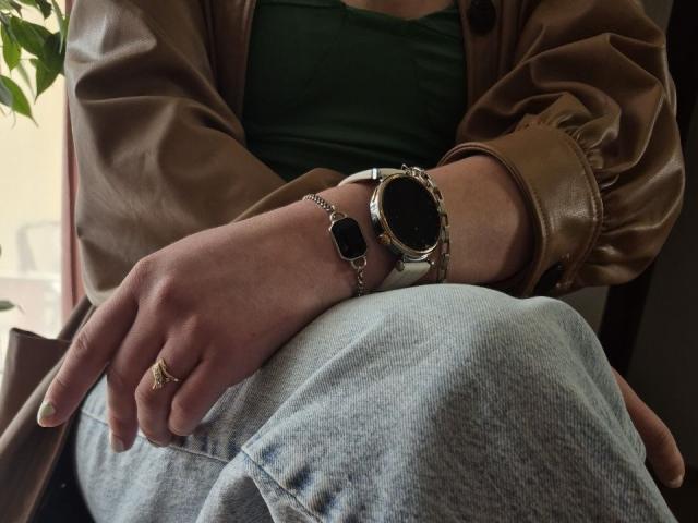 LoriEla trägt graue Jeans, ein grünes Oberteil, eine braune Lederjacke, eine schwarze Smartwatch, ein silbernes Armband und einen goldenen Ring. Ein Finger ist leicht sichtbar..