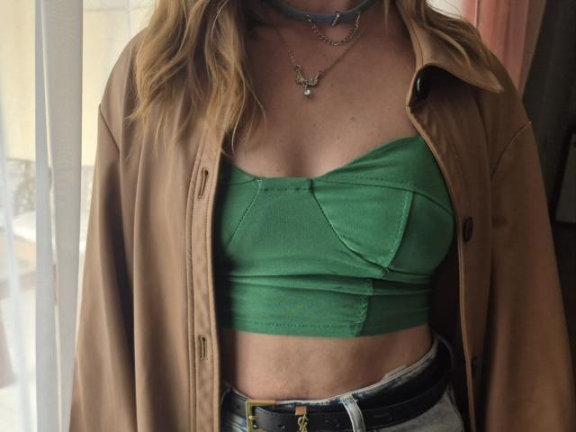 LoriEla con un top verde senza spalline, una giacca marrone e una cintura nera, che mostra il seno, i capelli biondi ondulati e due collane, in piedi di fronte a una finestra..