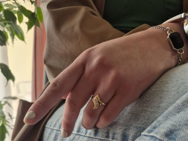 La main de LoriEla, avec une bague en or, un doigt pointé, un vernis à ongles blanc et une montre intelligente noire, portée sur un jean bleu. Une veste marron, une chemise verte et une plante sont visibles en arrière-plan..
