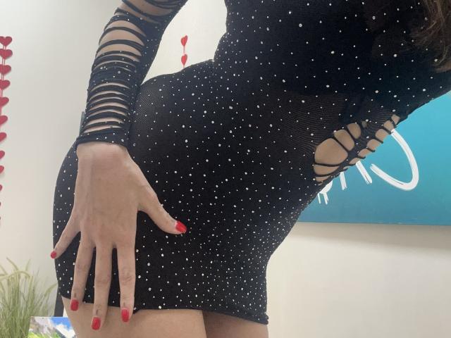 MoonLiza wearing a black, sparkly, cut-out dress with mesh sleeves, red nails, hand on curvy butt, no visible tits or pussy. Blue wall and red decorations in background.