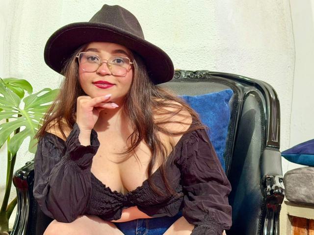 SamyMancini, con lunghi capelli castani, occhiali, cappello nero e un profondo scollo in una maglia nera a spalle scoperte, appoggia il mento sulla mano, mostrando il seno..