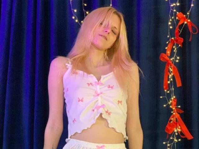 Dierdre Lorenzano mit blonden Haaren, die ein weißes Oberteil mit Blumenmuster und Rüschen sowie weiße Hosen trägt, steht vor einem blauen Vorhang mit Lichterketten und roten Schleifen..