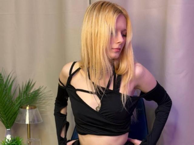 Dierdre Lorenzano a les cheveux blonds et porte un haut noir à fines bretelles, qui laisse entrevoir son ventre, avec de longues manches, révélant ses bras fins et pâles ainsi que sa petite poitrine. Elle regarde en bas, légèrement sur le côté..