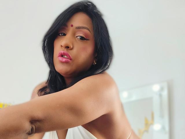 Nahirr has dark hair, pink lipstick, and a red bindi on her forehead. Her brown skin is smooth, and she's wearing a white top, showing her bare shoulders.