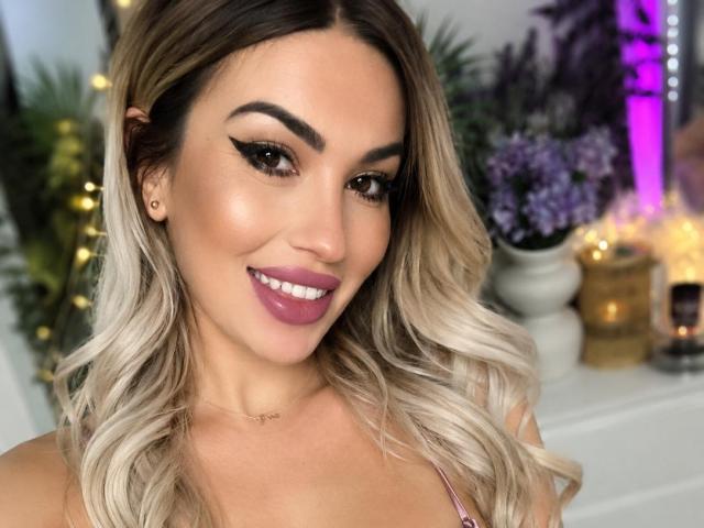 ZoeyMarie has blonde, wavy hair, brown eyes, thick black eyeliner, purple lipstick, and a cute smile. Her tits are visible, wearing a pink top, in a selfie.