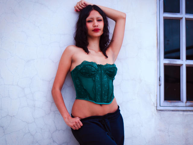 FreyaHommer with dark curly hair, wearing a green lace-up bustier, exposing her tits, and black pants, standing against a white cracked wall with a window on the right.