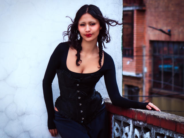 FreyaHommer with dark curly hair, wearing a black corset and long sleeves, leaning on a white and red brick wall, showing her tits, standing confidently with a slight smile.