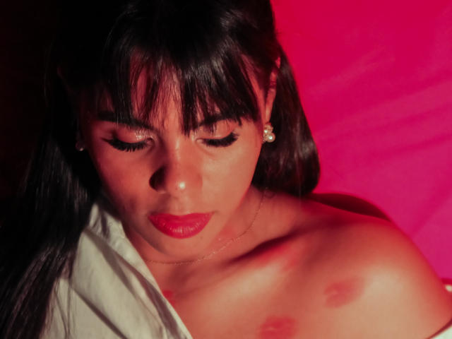 LissReyess a les cheveux foncés, du rouge à lèvres, et les yeux fermés. Elle est nue, avec des mamelons visibles, elle porte des boucles d'oreilles en diamants, et une chemise blanche recouvre partiellement ses épaules. Une lumière rouge met en valeur sa silhouette..