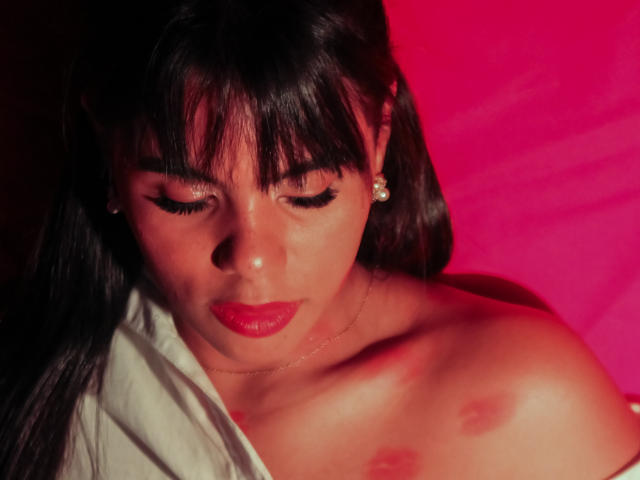 LissReyess, con capelli scuri, rossetto rosso e eyeliner, che guarda in basso, senza indumenti superiori, con piccoli capezzoli rotondi, indossa una collana e orecchini, su uno sfondo rosa..