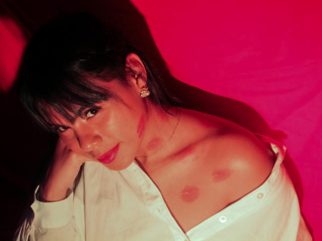LissReyess, con capelli scuri, frangia e orecchini, indossa una camicia bianca portata da una spalla, che mostra il seno, con rossetto rosso, su uno sfondo rosa, sorridendo..