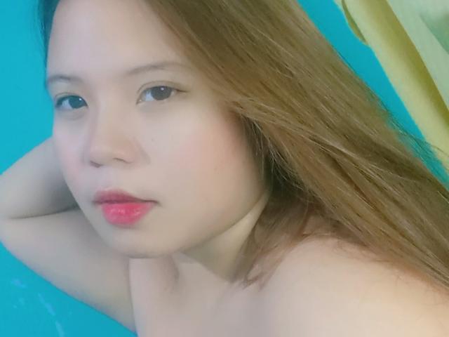 YannaDirtyOne with light skin, pink lipstick, and long brown hair, arms behind head, topless, looking at camera with a slightly pouty expression. Blue background.