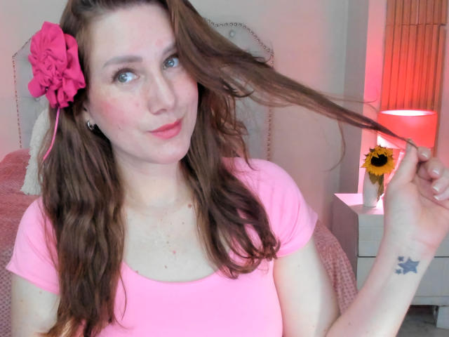 KatyaRogersX with pink flower in brown hair, pink shirt, holding hair, blue eyes, slight smile, small star tattoo on right wrist, white bedroom background, red lamp.