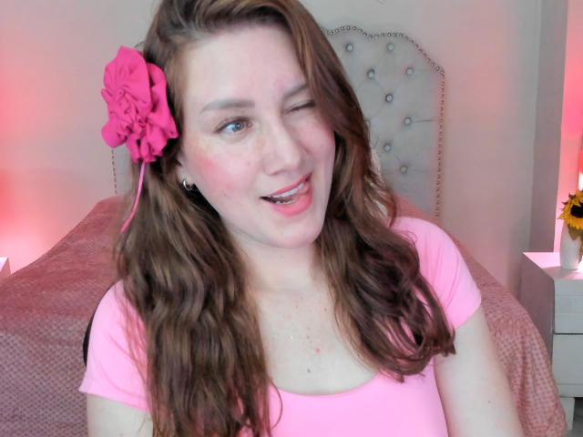 KatyaRogersX, clignant des yeux et la langue rose sortant, a une fleur rose dans ses cheveux bruns ondulés, porte un haut rose, a des taches de rousseur et une peau claire, dans une chambre avec une tête de lit capitonnée..