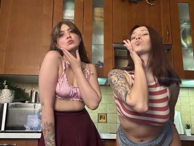 HotCaramelsTrio avec des cheveux bruns, un haut rose à volants et une jupe marron, et HotCaramelsTrio avec des cheveux roux-bruns, un crop top rayé et un short en jean, se faisant des signes de la main. Les deux ont des tatouages..