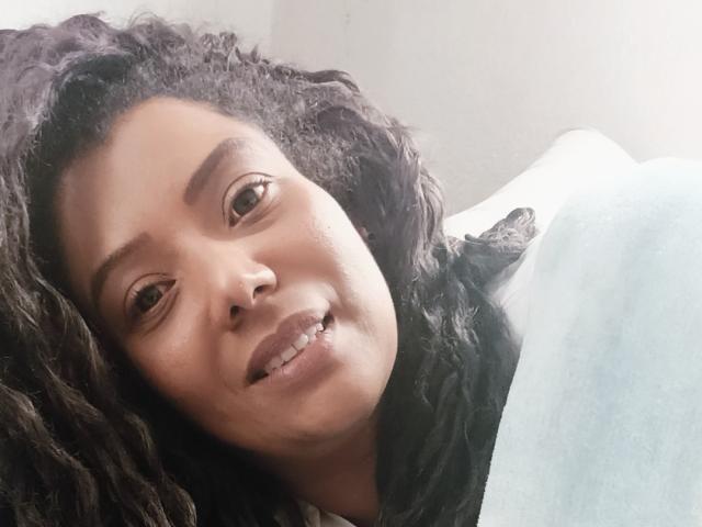 KenyaKarath hat lockiges, schwarzes Haar, einen Nasenring und ein leichtes Lächeln mit einer kleinen Lücke zwischen den Zähnen. Sie wirkt entspannt, während sie unter einer blauen Decke liegt..