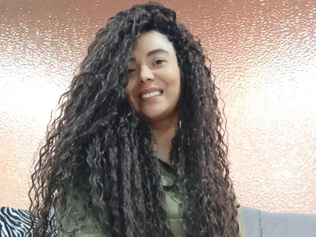 KenyaKarath, com cabelos longos, cacheados e castanho-escuros, sorrindo, vestindo uma camisa verde, tem pele marrom-média e está em pé em frente a uma parede rosa texturizada, mostrando os dentes..