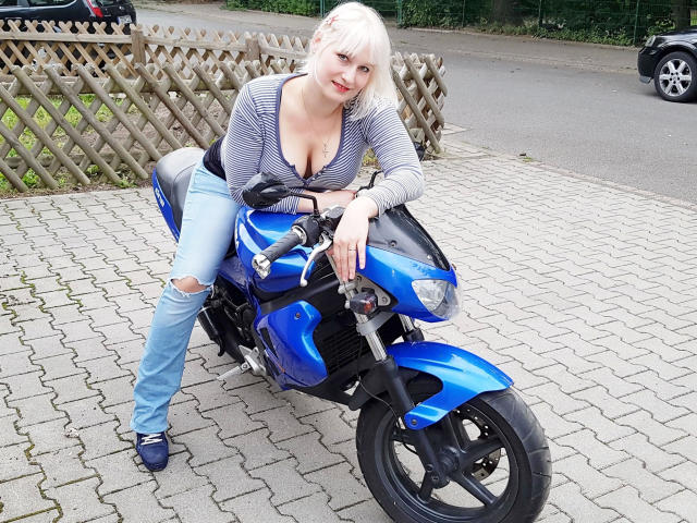 Ginger-Babe-hot with blonde hair, wearing a striped top and ripped jeans, leaning on a blue motorcycle, showing her tits, with a sexy smile, and blue shoes.