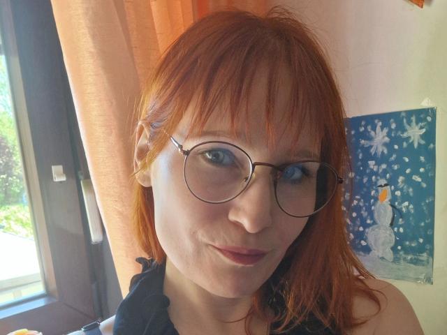 Ginger-Babe-hot with red hair, glasses, and a slight smile, showing her cute face and small tits, wearing a black top, in a sunlit room.