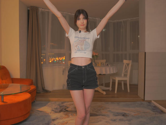 ElmaFasula with arms raised, wearing a white crop top and dark denim shorts, standing in a room with orange furniture and a blue rug. Her tits are small.