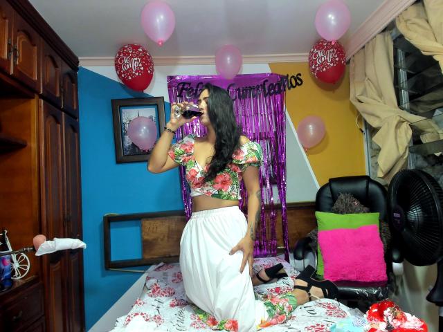 AfroditaHan with floral crop top, white skirt, and black heels, sipping from a bottle, kneeling on a bed, surrounded by pink and red balloons in a colorful room.